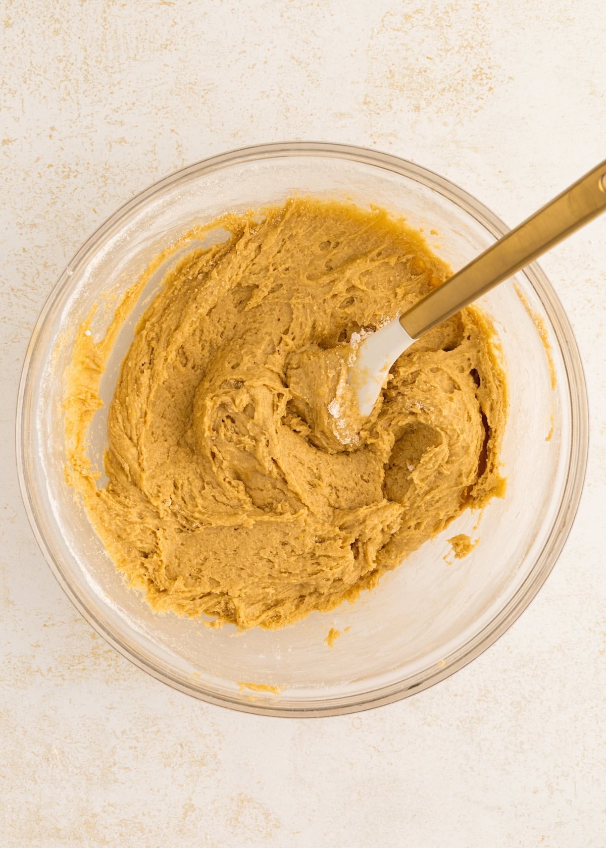 A glass bowl of brown cookie dough with a spatula on a light surface.