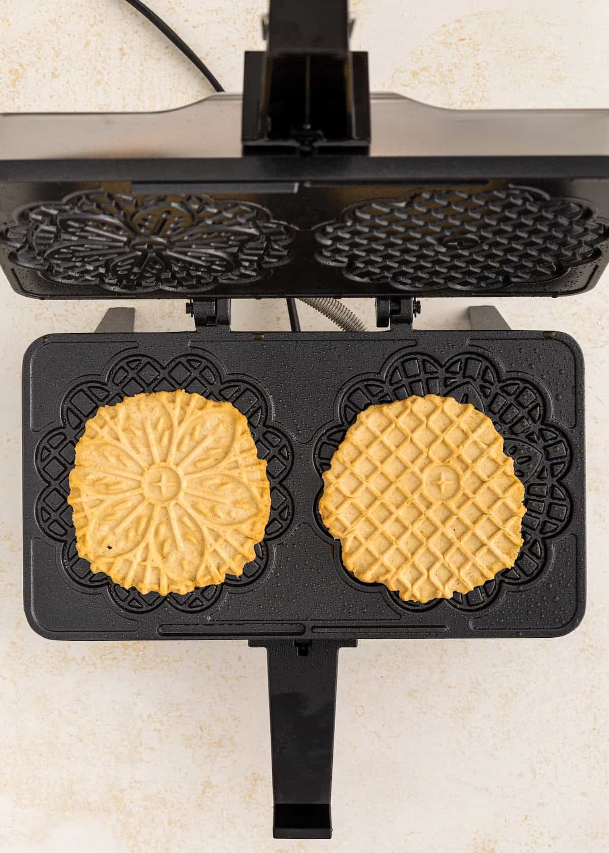 Two round pizzelle cookies cooking in a black pizzelle maker, viewed from above.