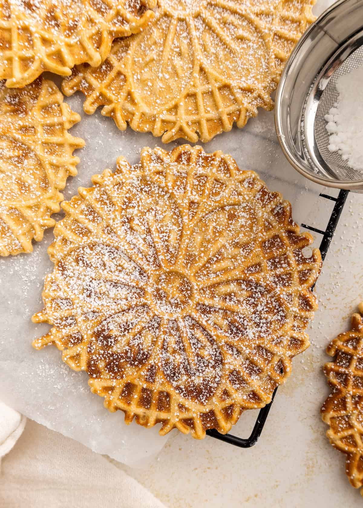 Round pizzelle cookies dusted with powdered sugar on a wire rack and parchment paper.