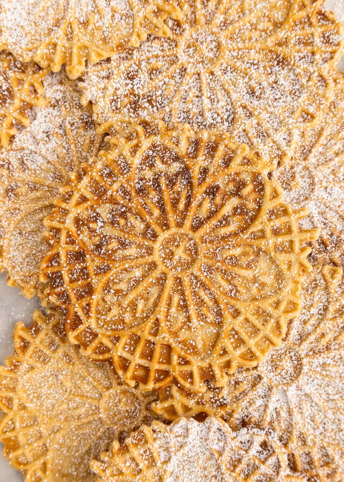 Golden, round pizzelle cookies dusted with powdered sugar, overlapping each other.