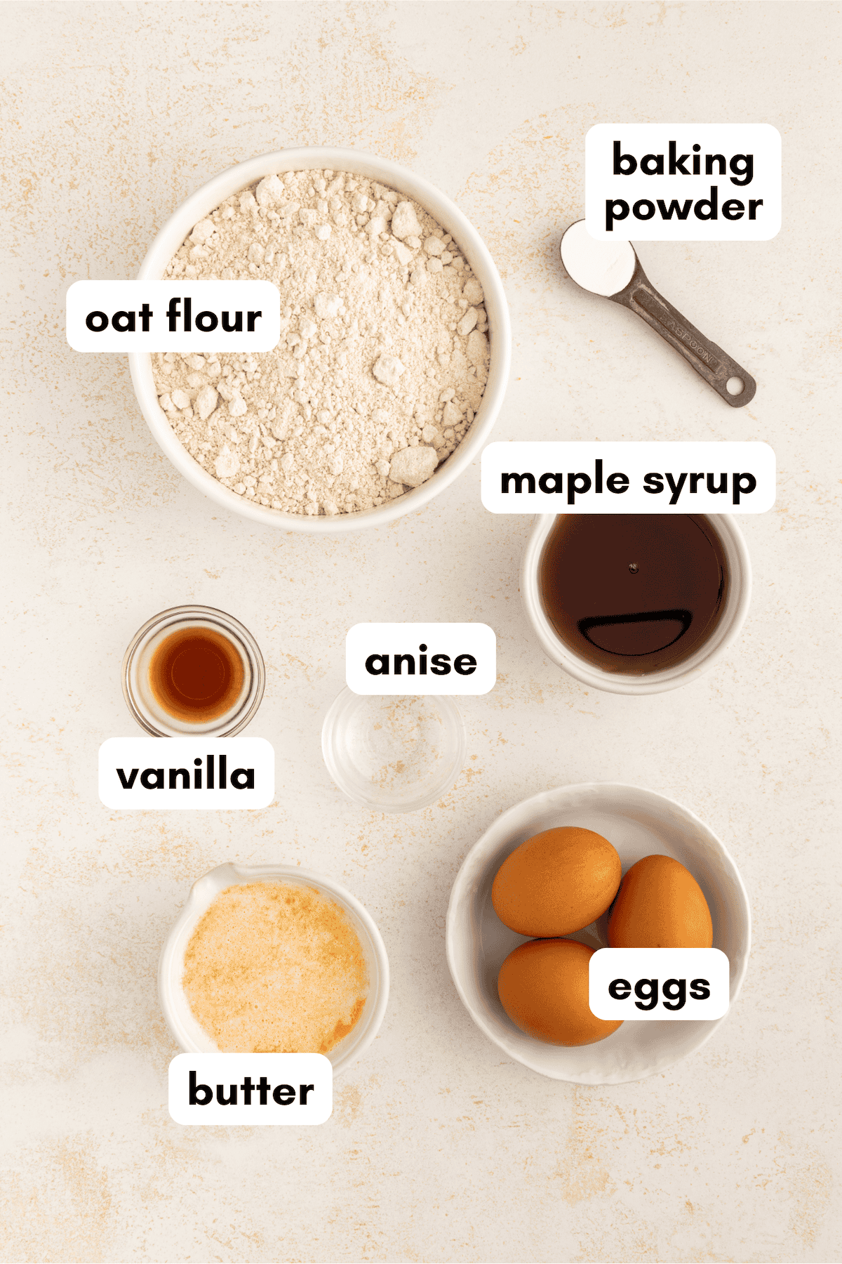 Baking ingredients on a light surface: oat flour, baking powder, eggs, butter, anise, vanilla, syrup.