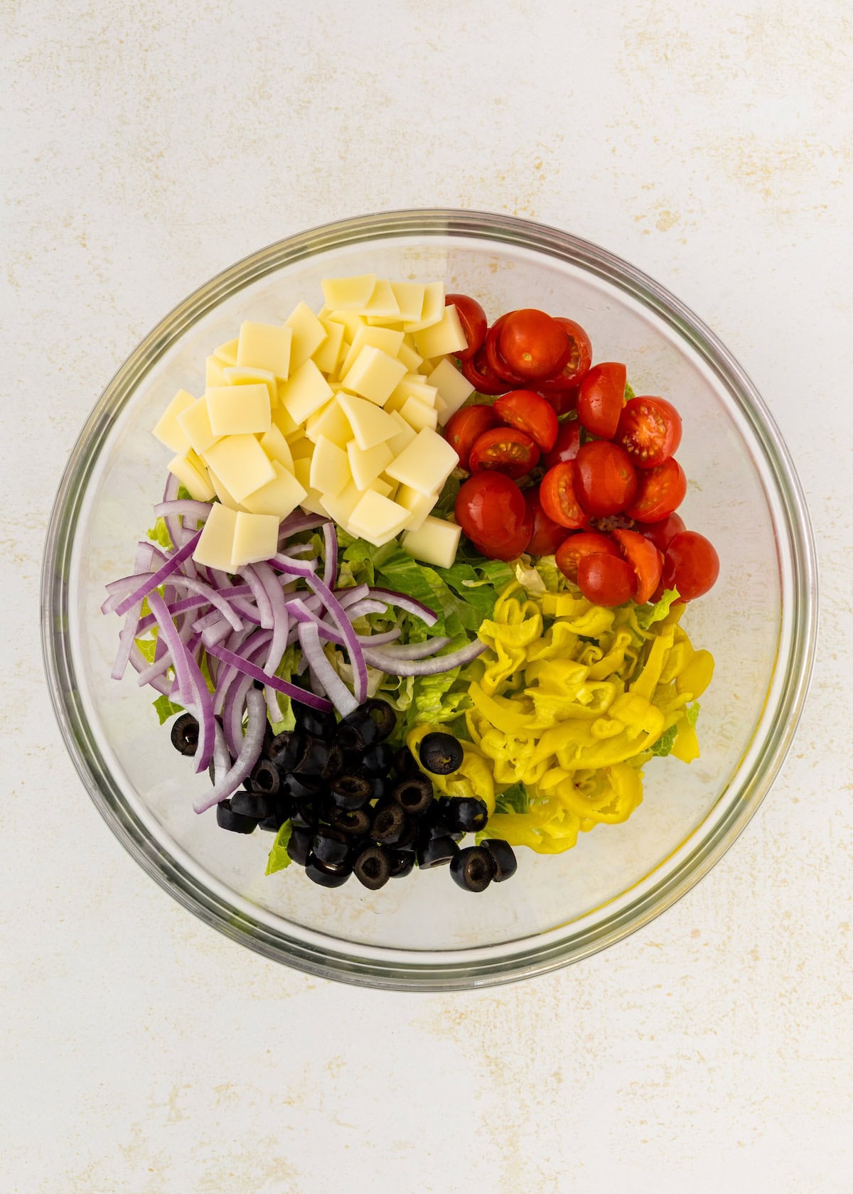 A bowl with lettuce, cheese cubes, tomatoes, onions, olives, and banana peppers.