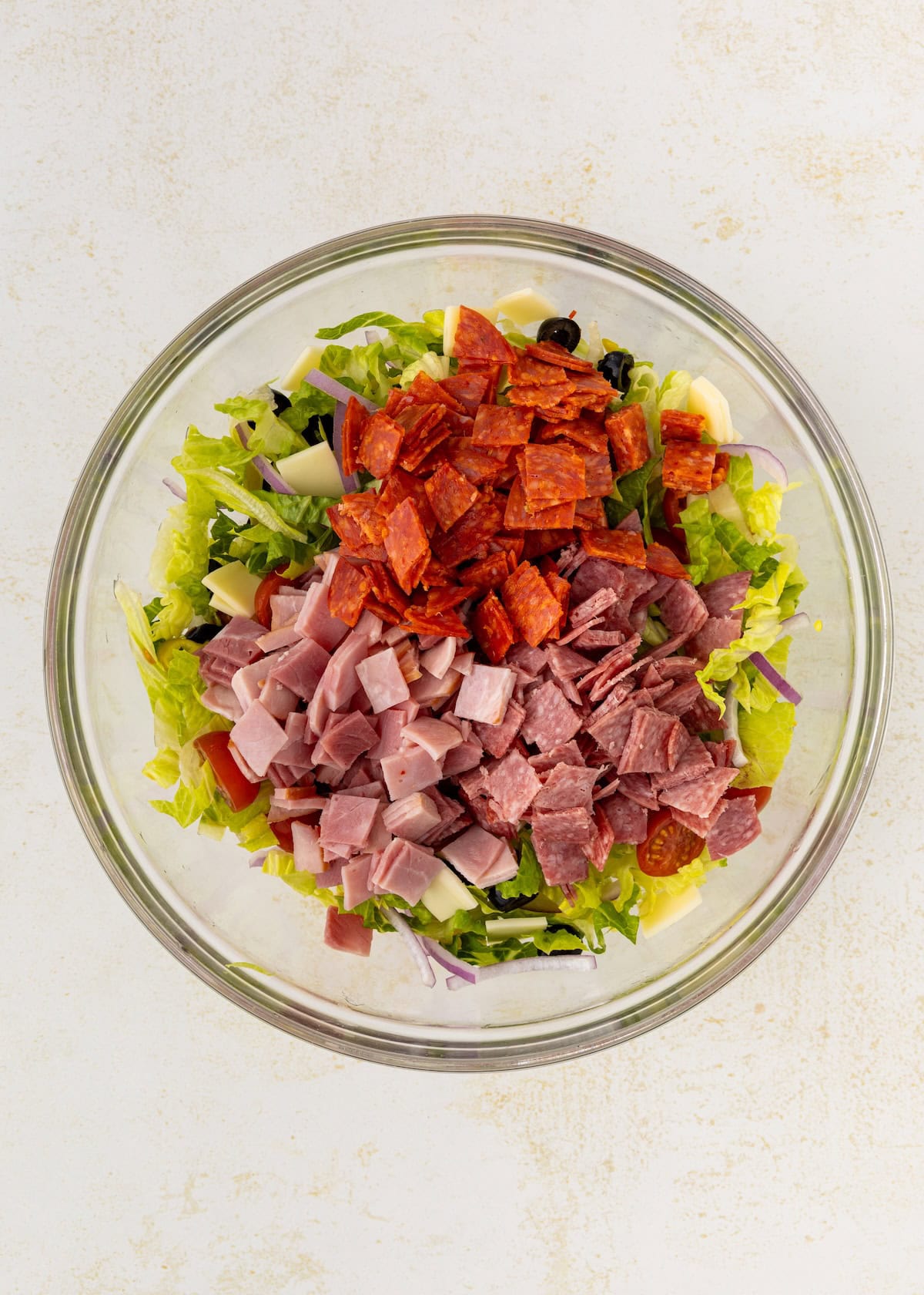 A glass bowl of chopped salad topped with ham, salami, and pepperoni slices.