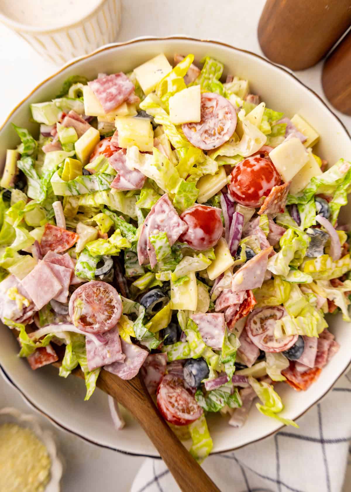 Chopped salad with lettuce, tomatoes, olives, cheese, and meats in a white bowl with a spoon.