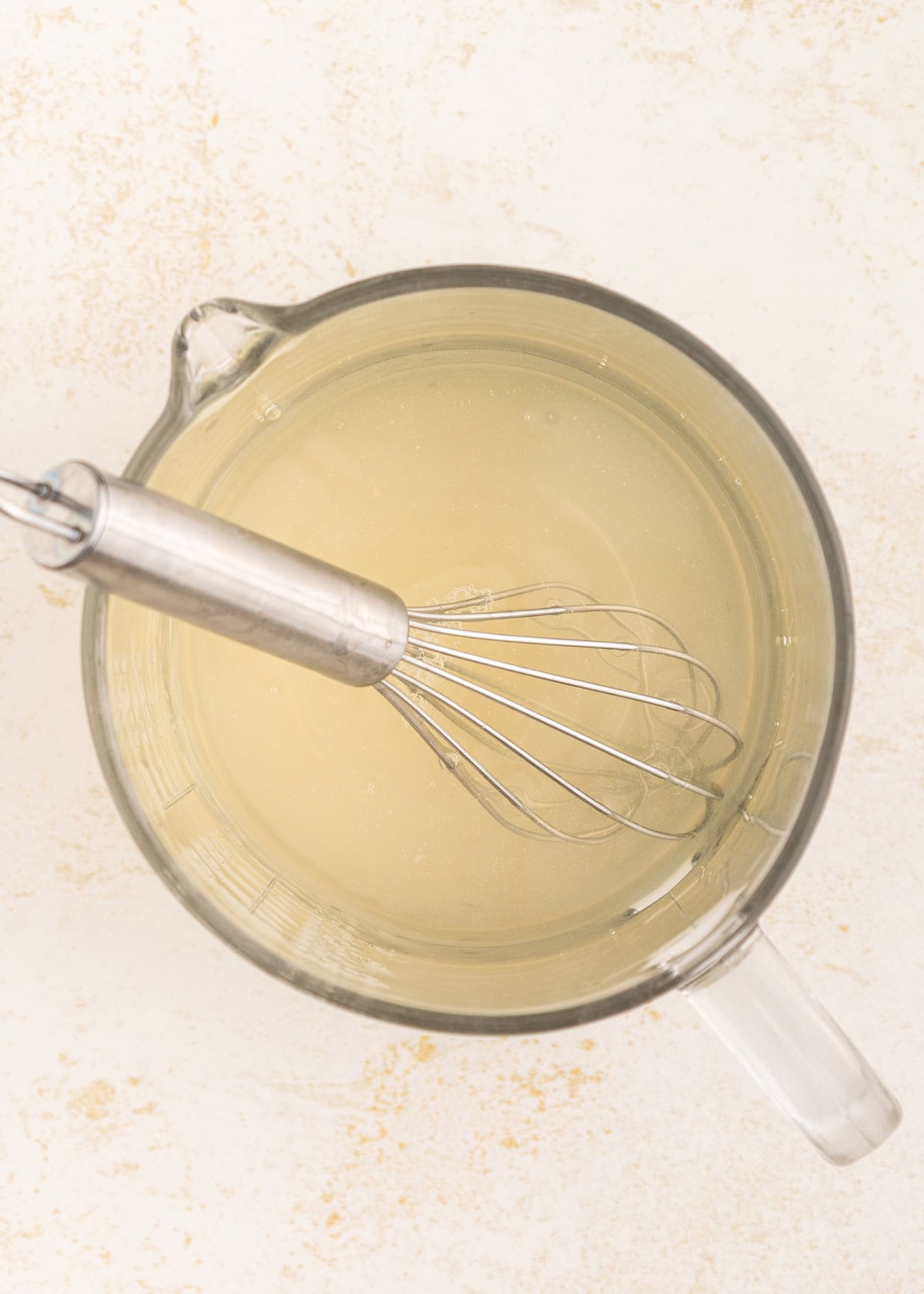 A metal whisk in a glass measuring cup of clear liquid on a light surface.