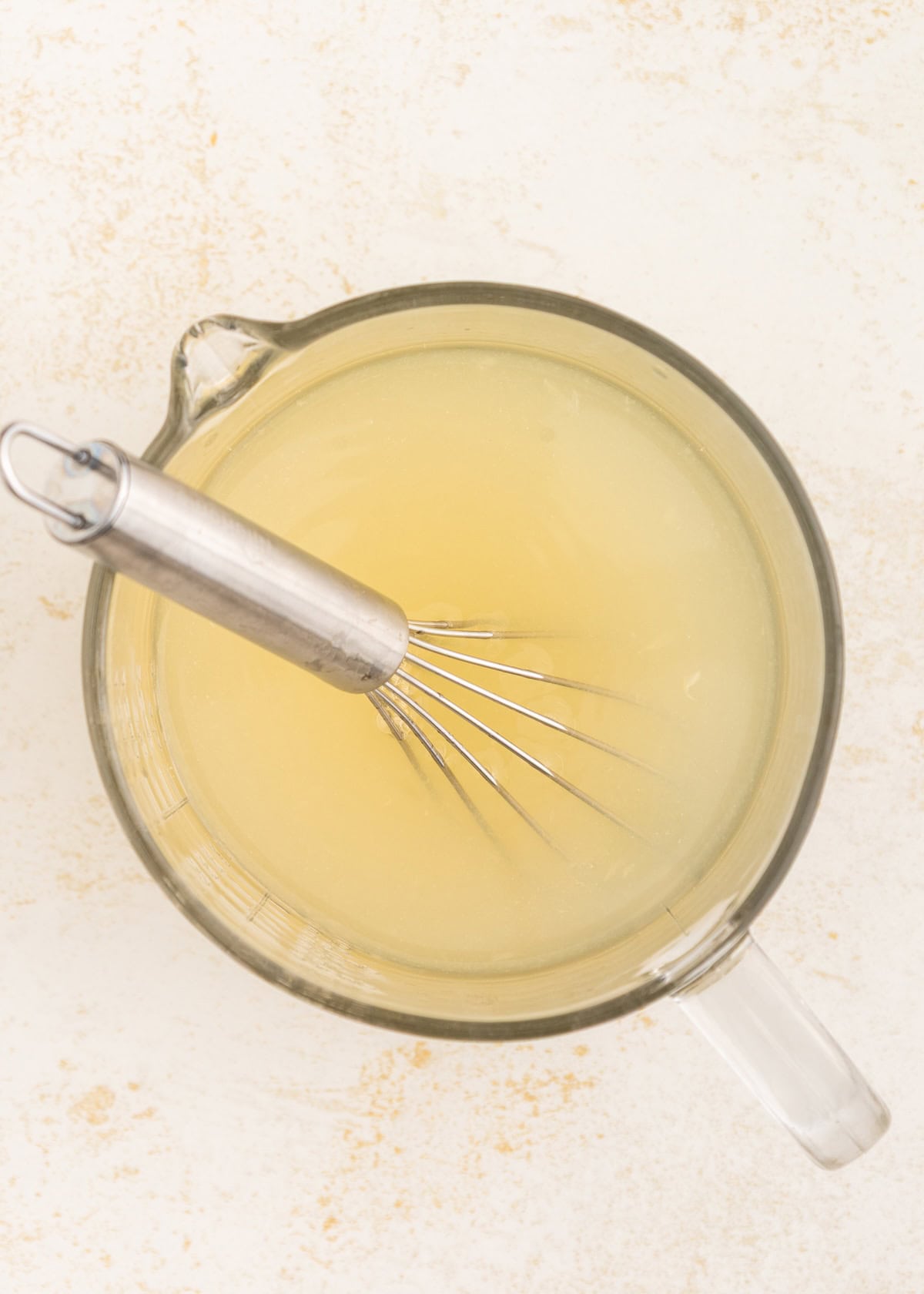 A metal whisk in a glass measuring cup filled with a pale yellow liquid.