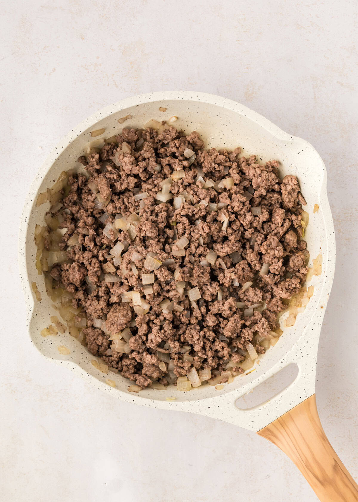 Ground beef and diced onions being cooked in a white skillet with a wooden handle.