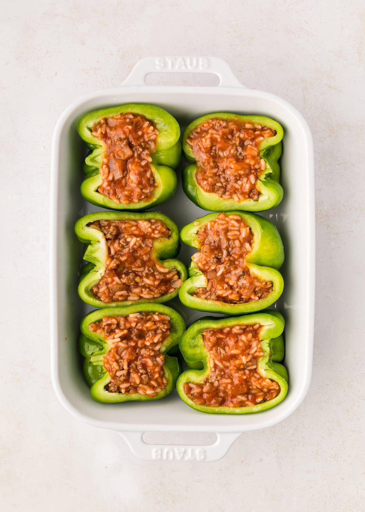 Six green bell peppers halved and filled with a red meat and rice mixture in a white baking dish.