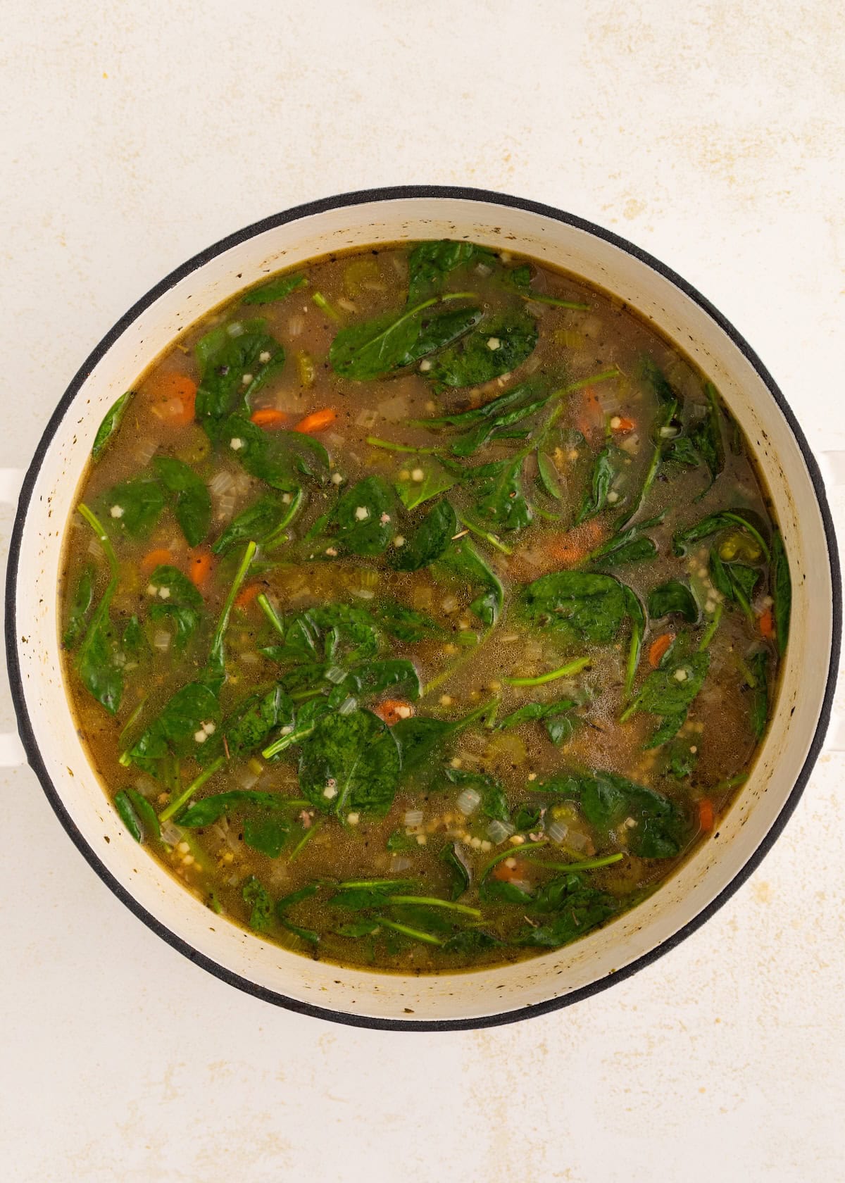 A pot of Italian Wedding Soup with spinach, carrots, and broth on a white surface.