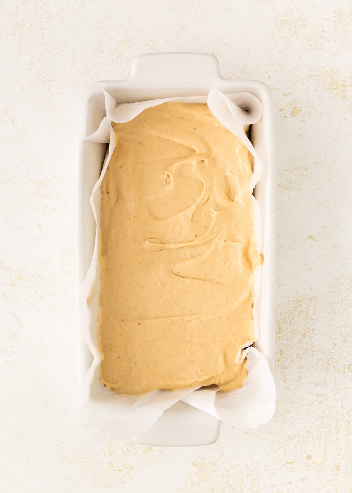 Loaf pan lined with parchment paper filled with smooth, light brown batter, overhead view.
