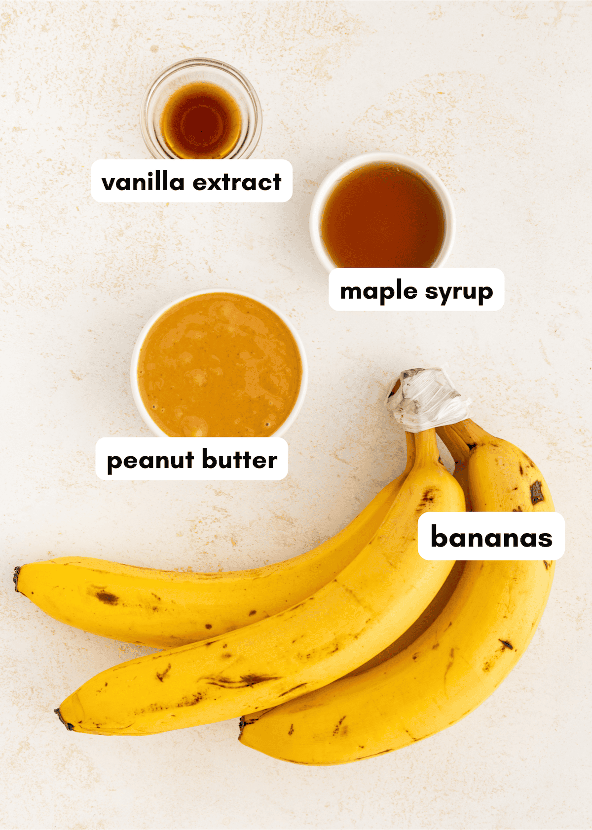 Four bananas, a bowl of peanut butter, maple syrup, and vanilla extract on a white surface.