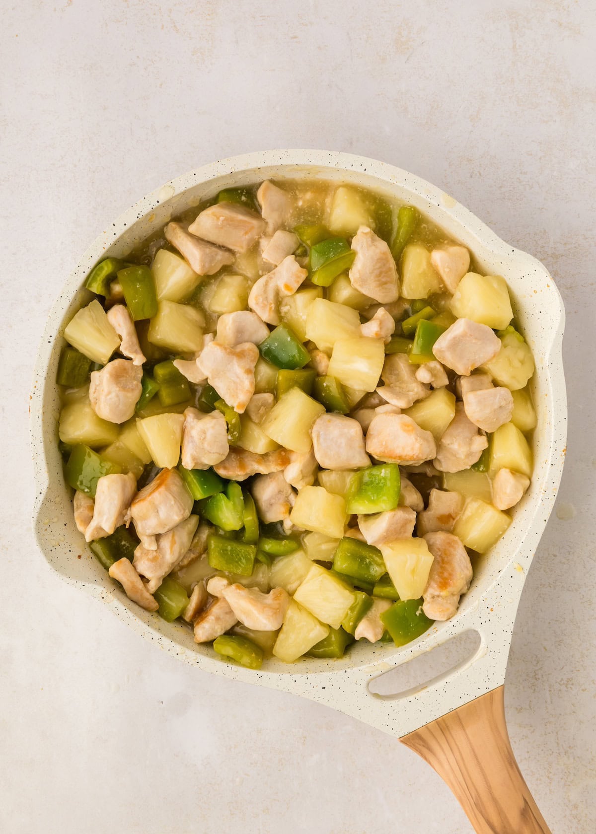 A skillet with chicken, pineapple chunks, and green bell peppers in a light sauce.