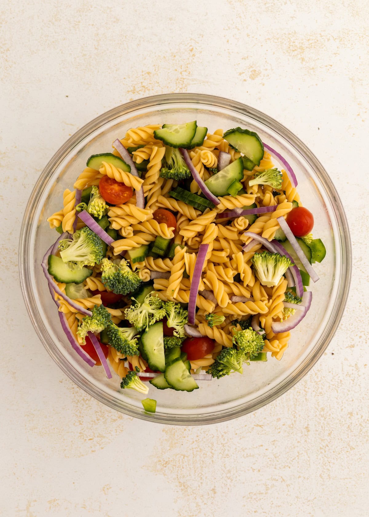 A glass bowl of protein pasta salad with broccoli, cucumber, tomato, and red onion.