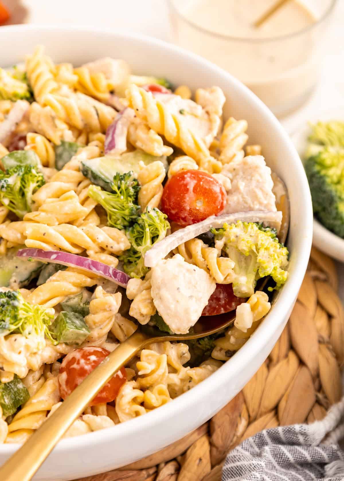 A bowl of protein pasta salad with chicken, broccoli, tomatoes, and red onion, plus a gold fork.