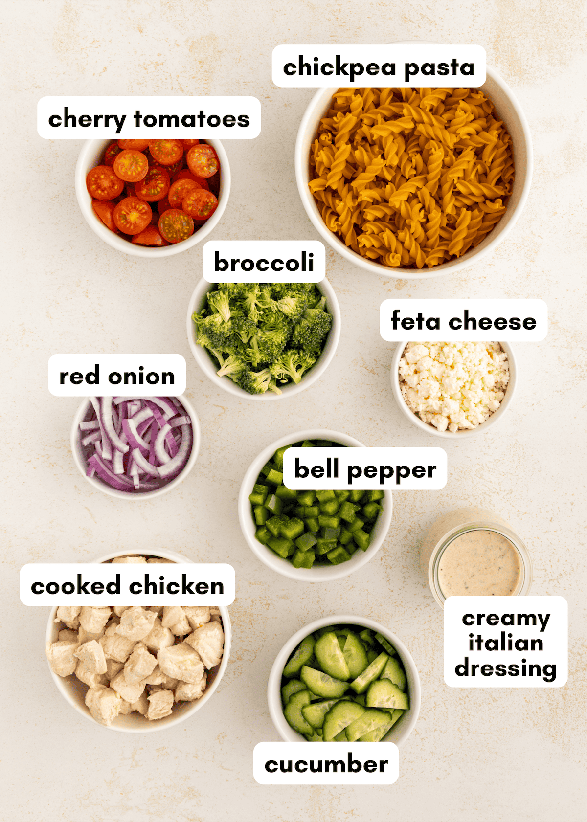Protein pasta salad bowls with veggies, feta, chicken, and creamy Italian dressing on a counter.