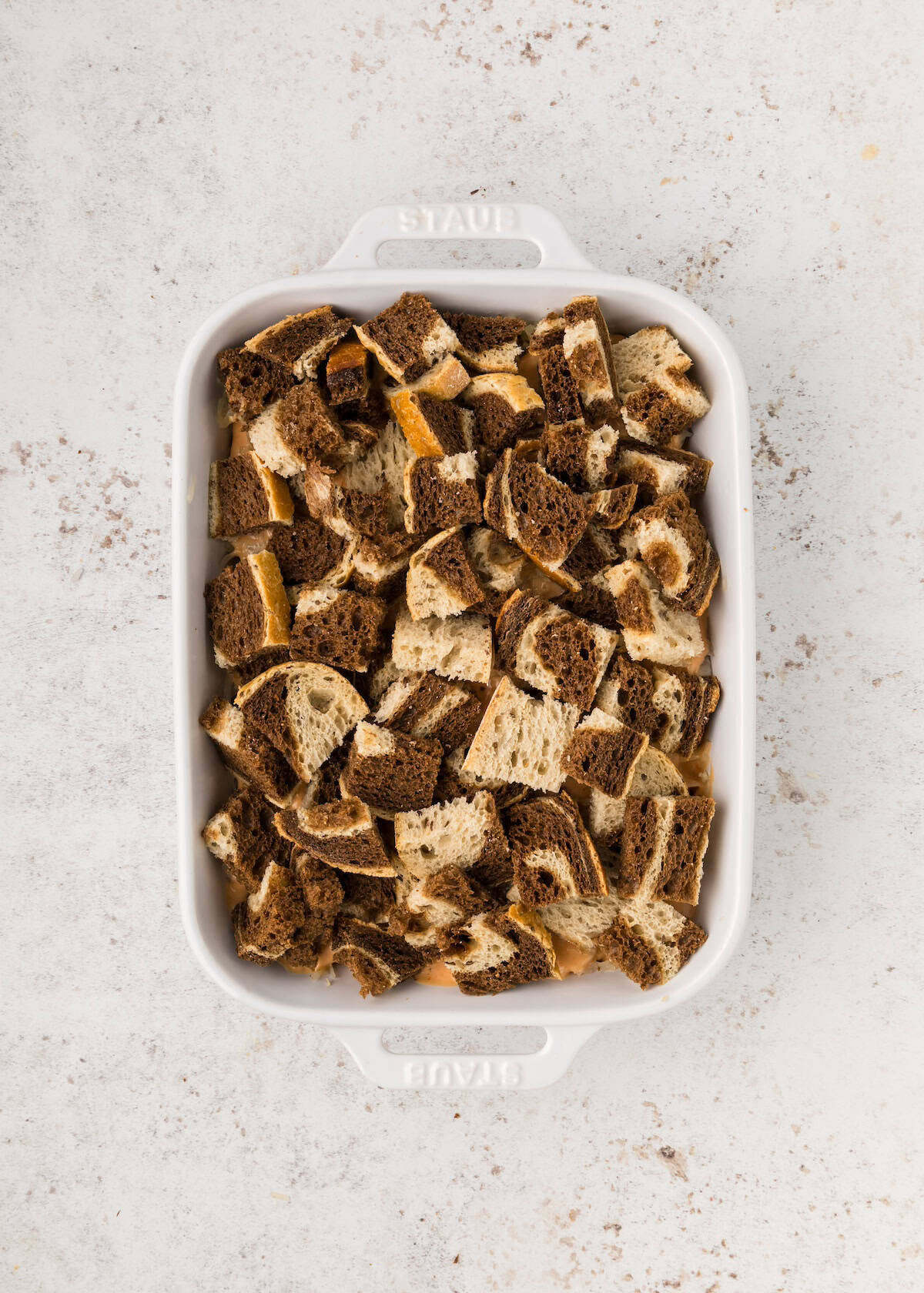Cubes of brown and white bread in a white rectangular baking dish on a light surface.
