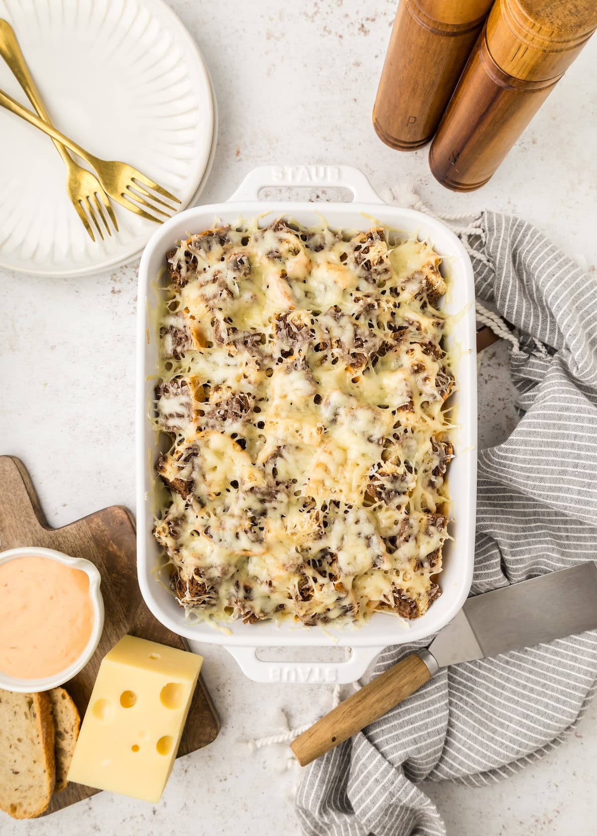A baked casserole topped with melted cheese in a white dish, beside plates and utensils.
