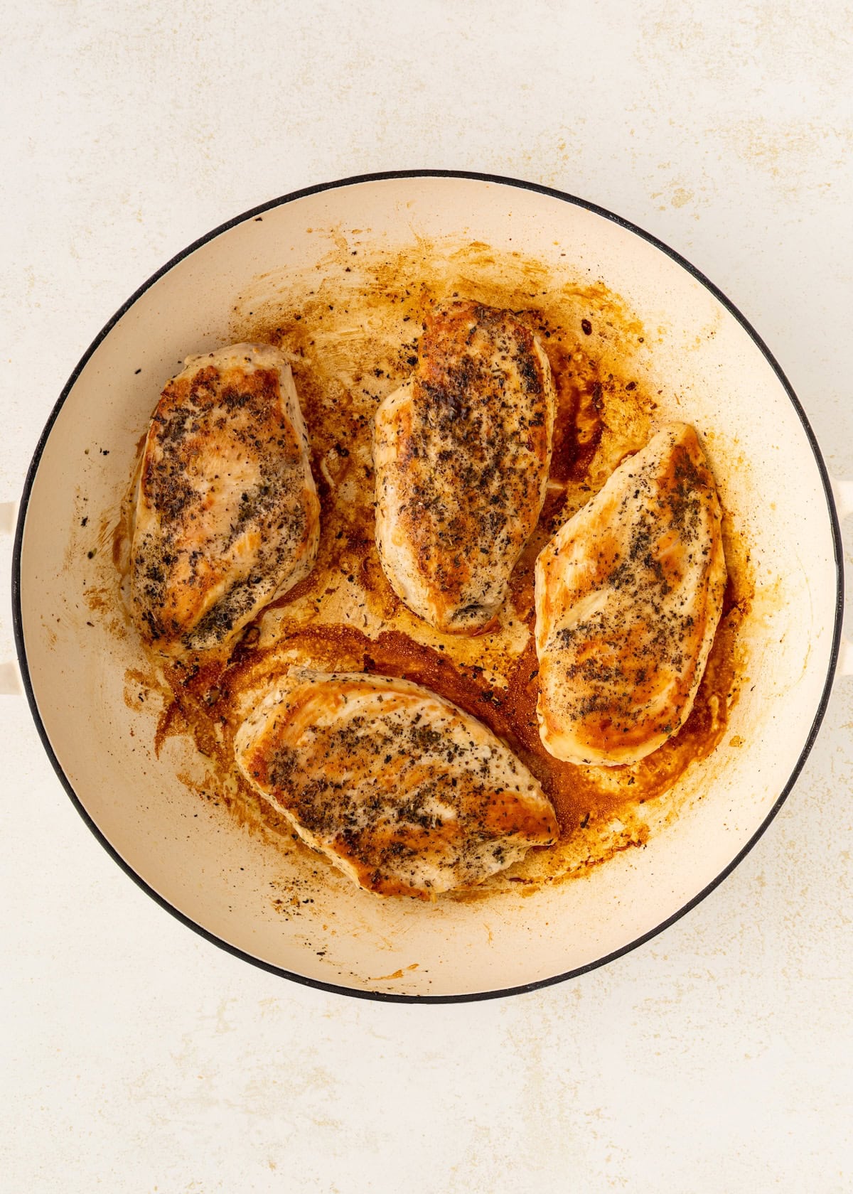 Four seasoned, cooked chicken breasts in a round white skillet on a light surface.