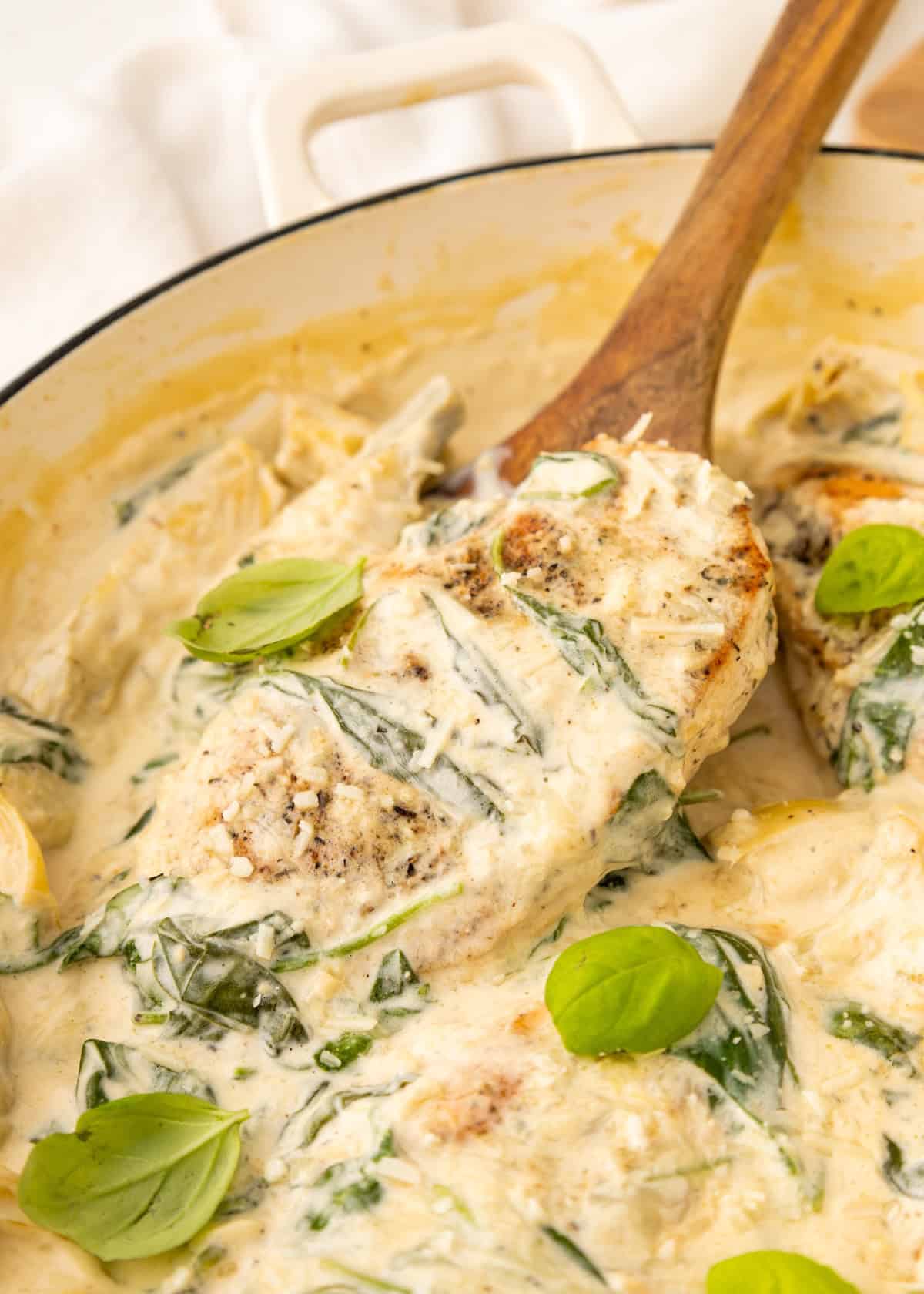 Creamy chicken breast with spinach and basil leaves in a skillet, served with a wooden spoon.