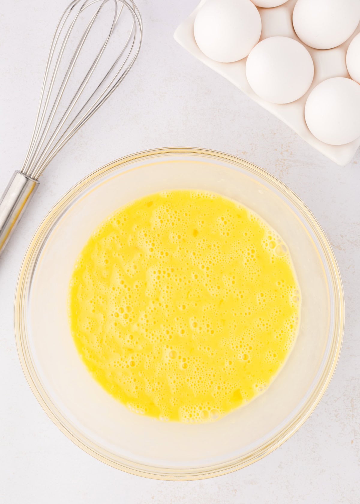 A bowl of beaten eggs next to a whisk and a carton of whole eggs on a white surface.