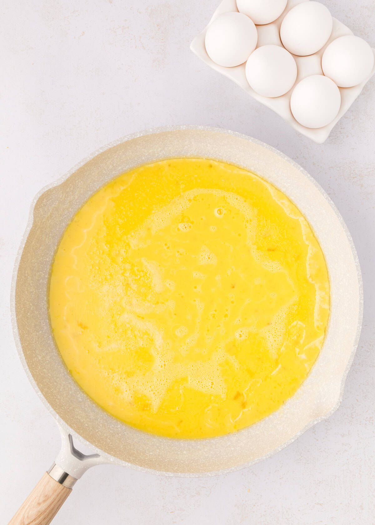 Pan with beaten eggs cooking, next to a carton holding six whole eggs on a light surface.
