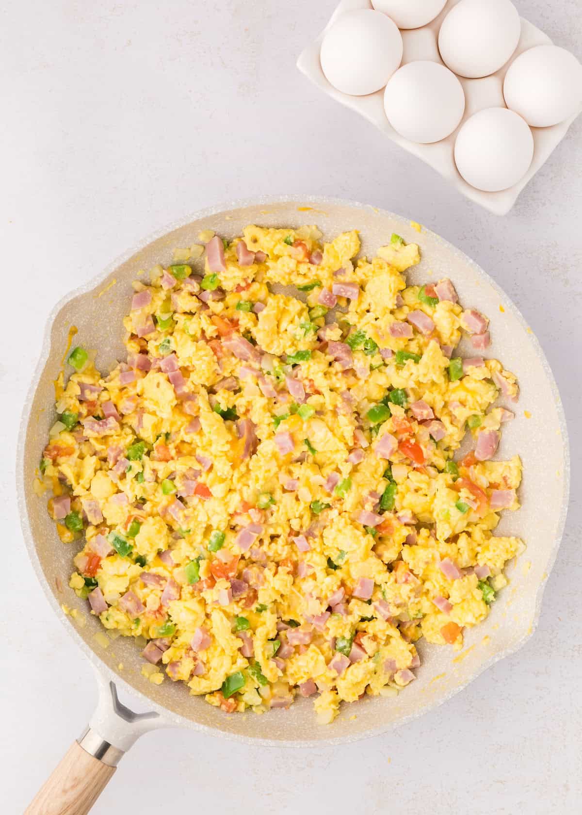Scrambled eggs with ham and vegetables in a pan, with whole eggs in a carton nearby.