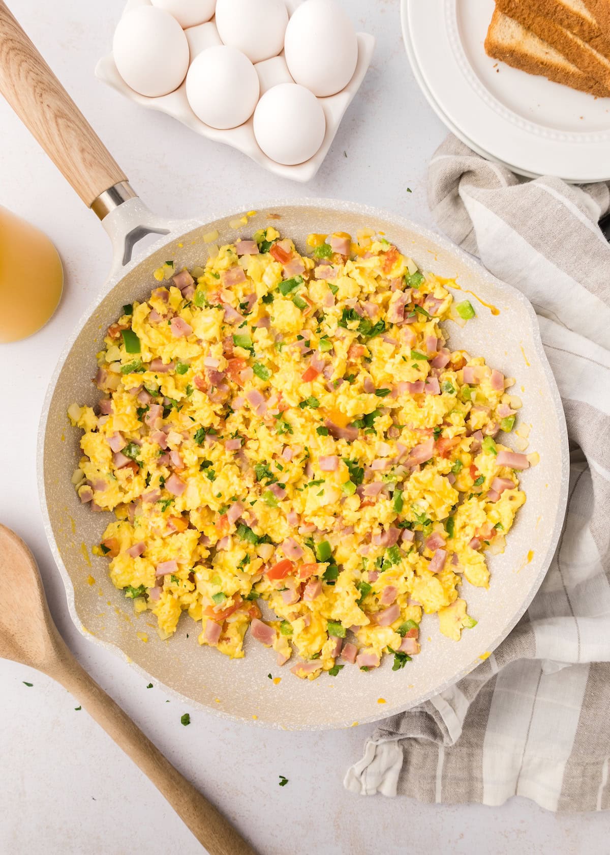 Pan of scrambled eggs with diced ham, tomatoes, and herbs, next to eggs and toast.
