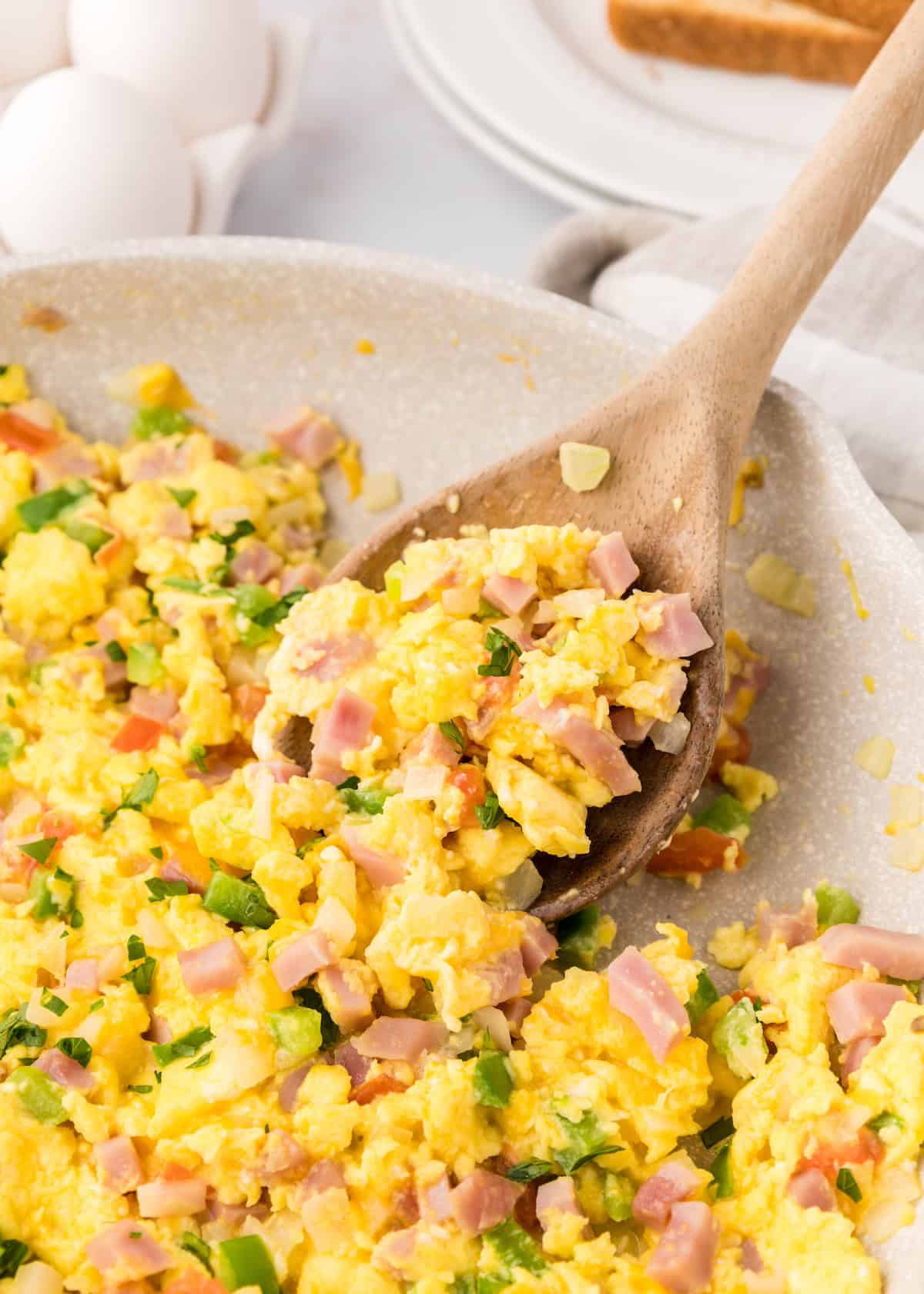 Scrambled eggs with diced ham and green peppers in a pan, stirred with a wooden spoon.