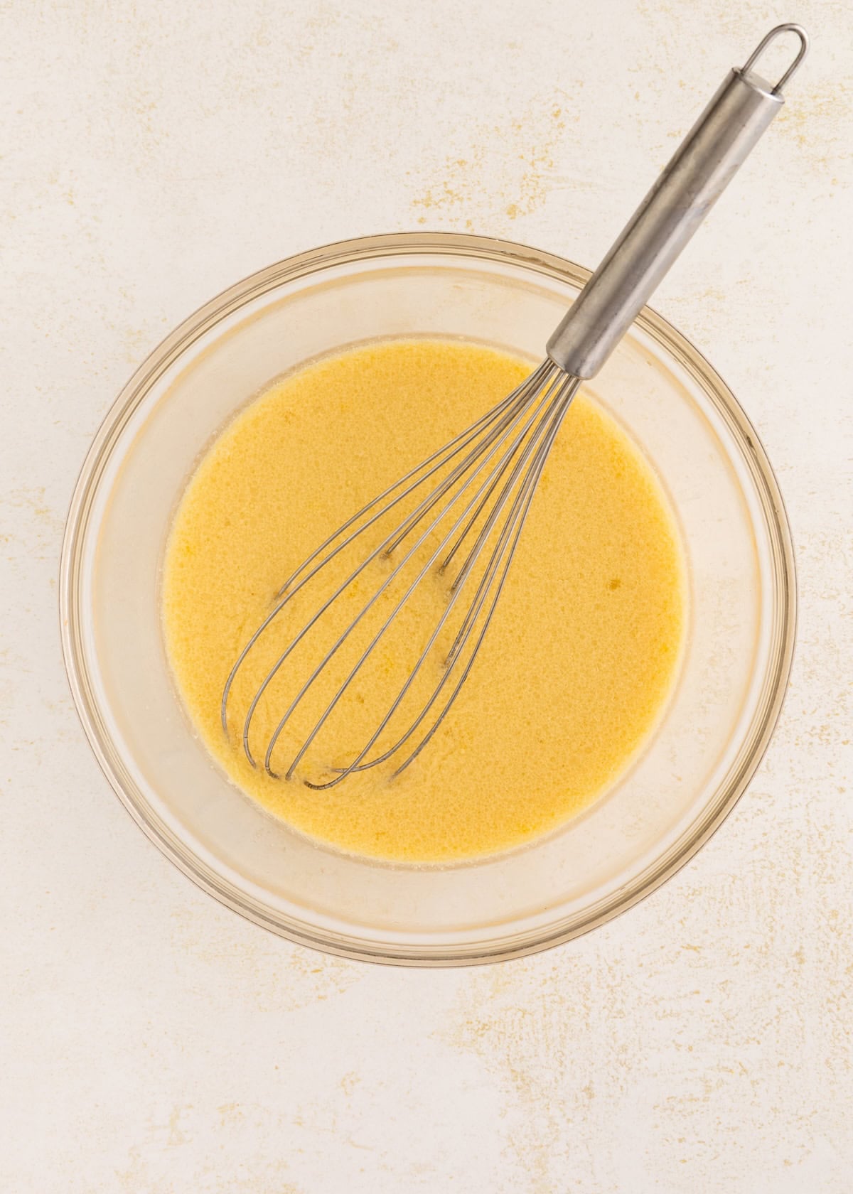 A whisk rests in a bowl filled with a yellow liquid mixture on a light surface.