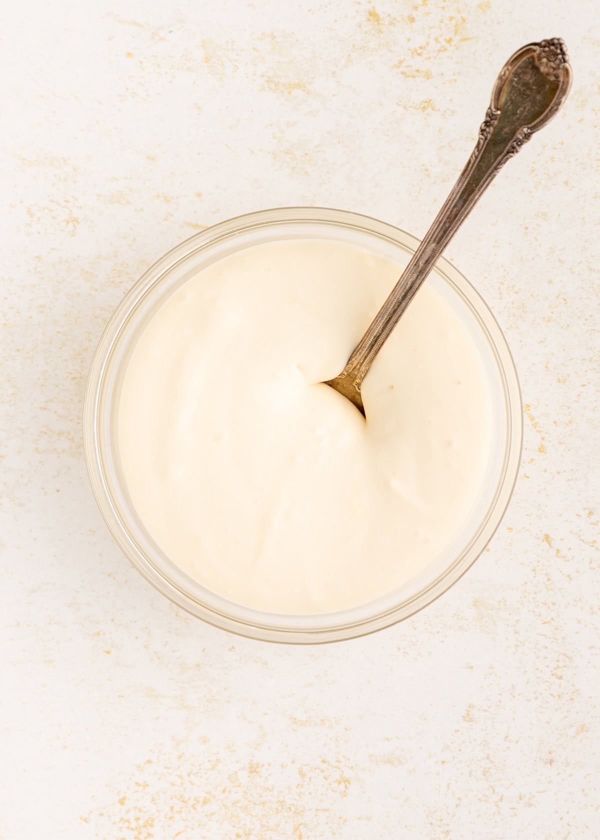 A glass bowl of creamy white sauce with a metal spoon on a light surface.
