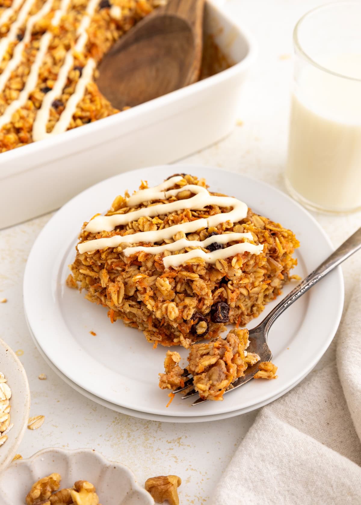 A slice of baked oatmeal with white icing drizzle on a plate, next to a glass of milk.