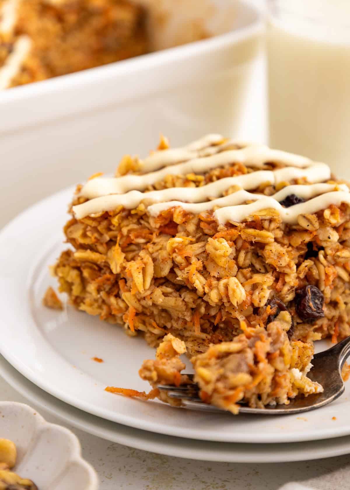 A slice of baked oatmeal with icing drizzle on a white plate, served with a spoon.