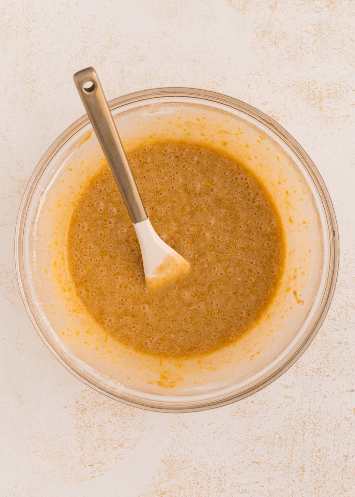 A glass bowl of brown batter with a white spatula on a light surface.