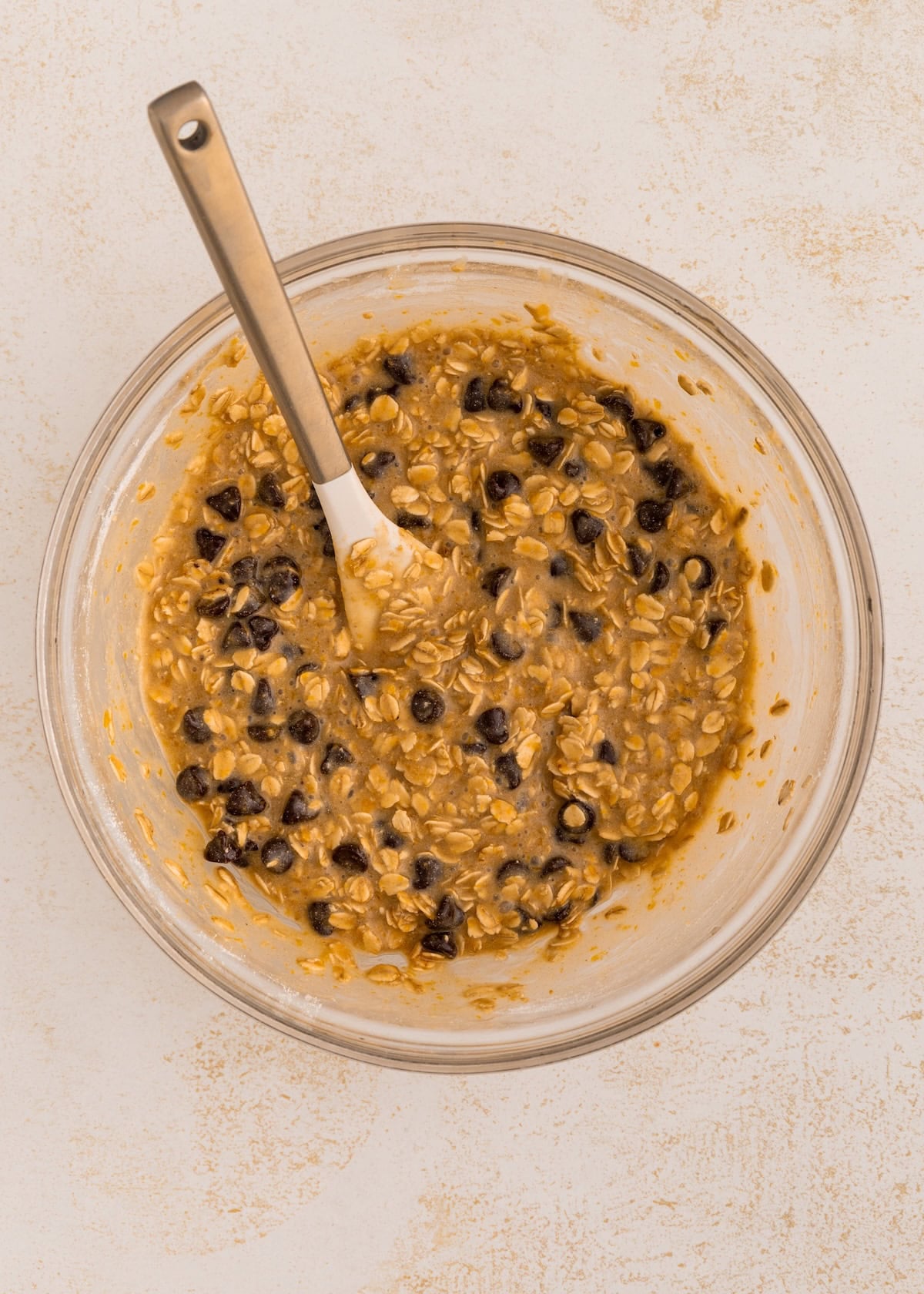 A glass bowl with oatmeal cookie dough and chocolate chips, stirred with a metal spoon.