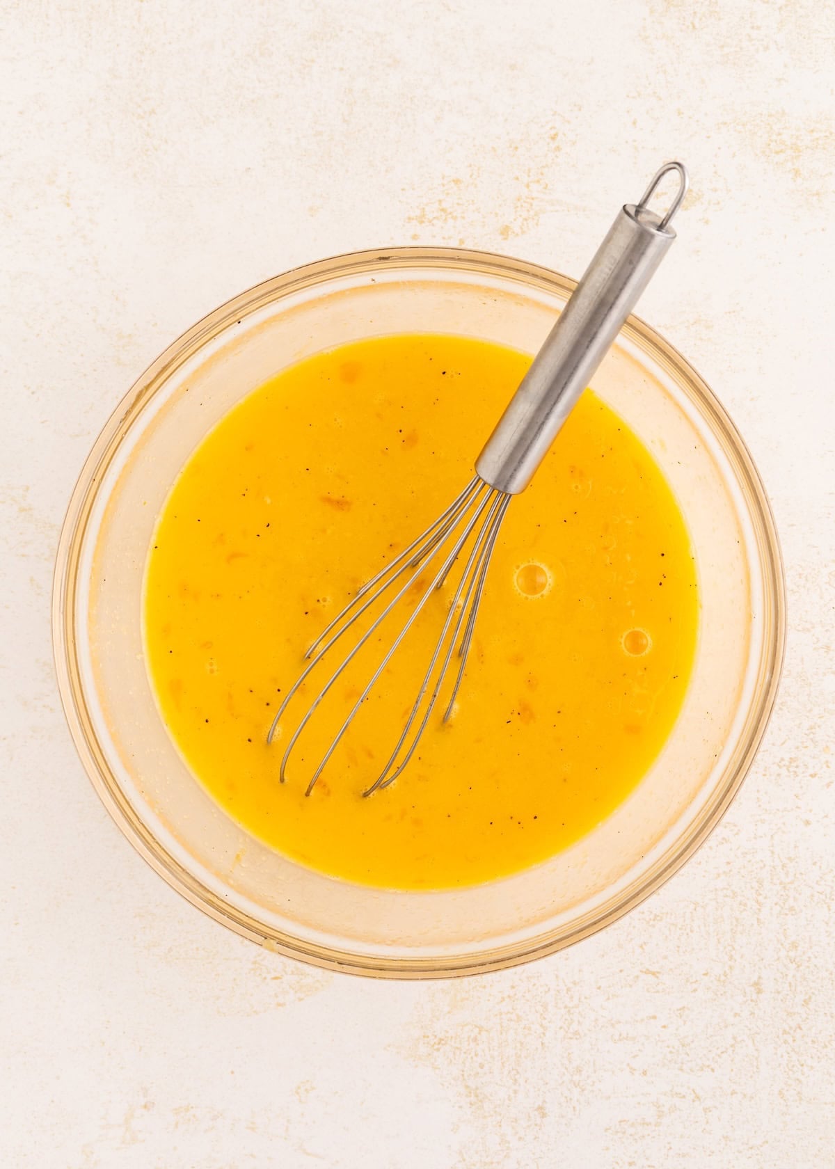 A glass bowl of beaten eggs with a metal whisk on a light-colored surface.