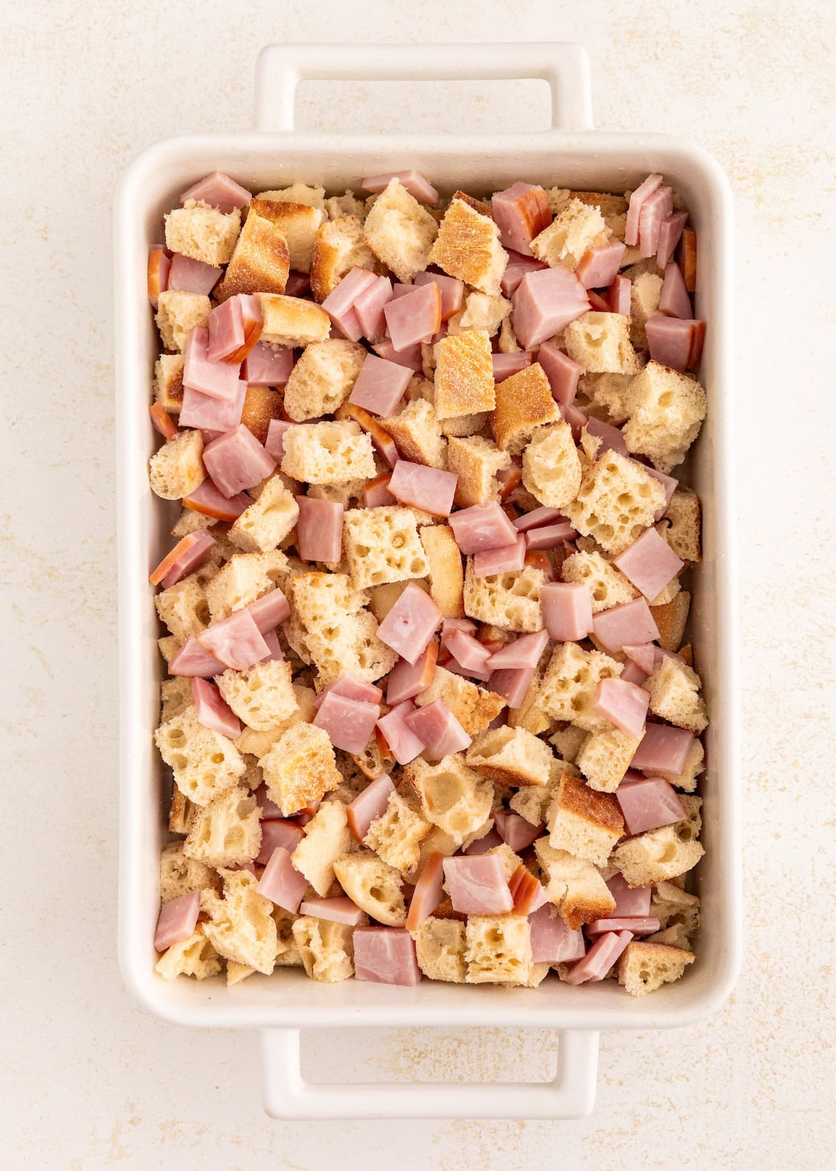 Cubed bread and diced ham in a white rectangular baking dish on a light surface.