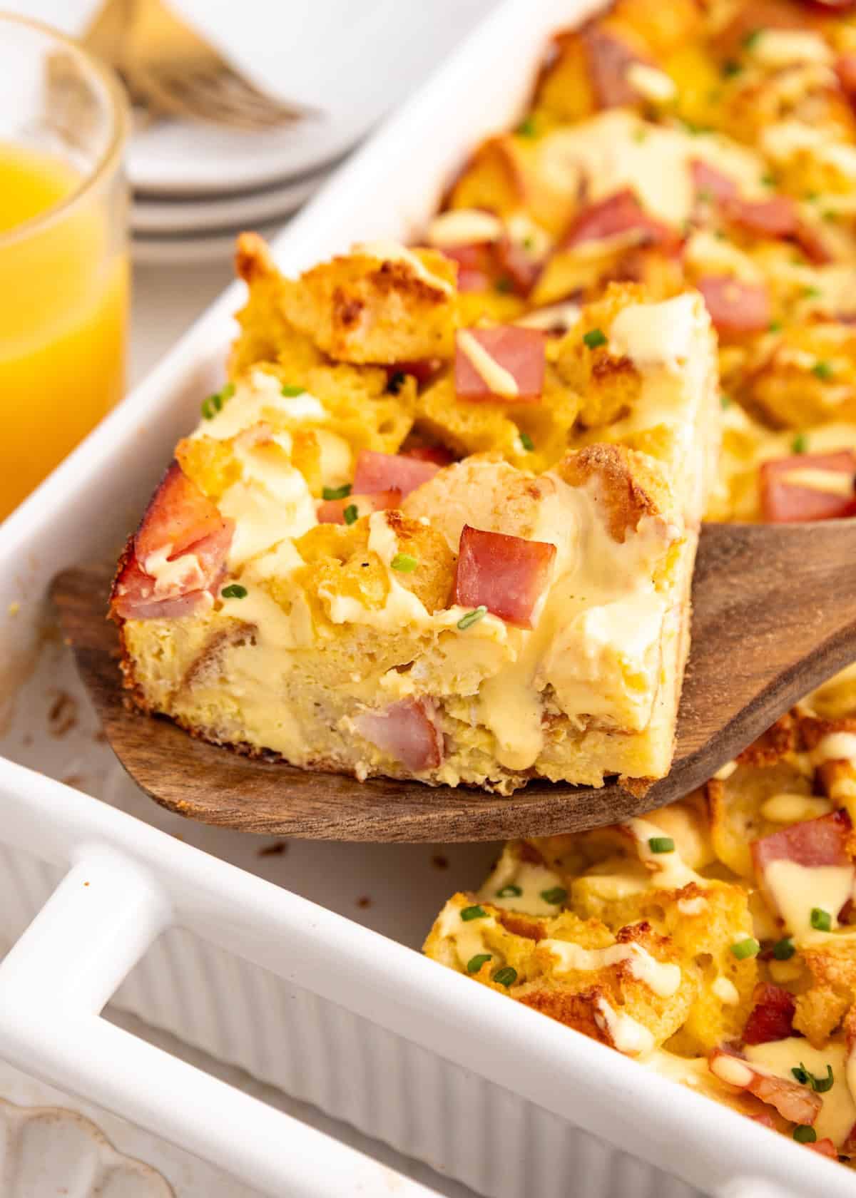 A spatula lifts a cheesy ham and egg breakfast casserole from a white baking dish.