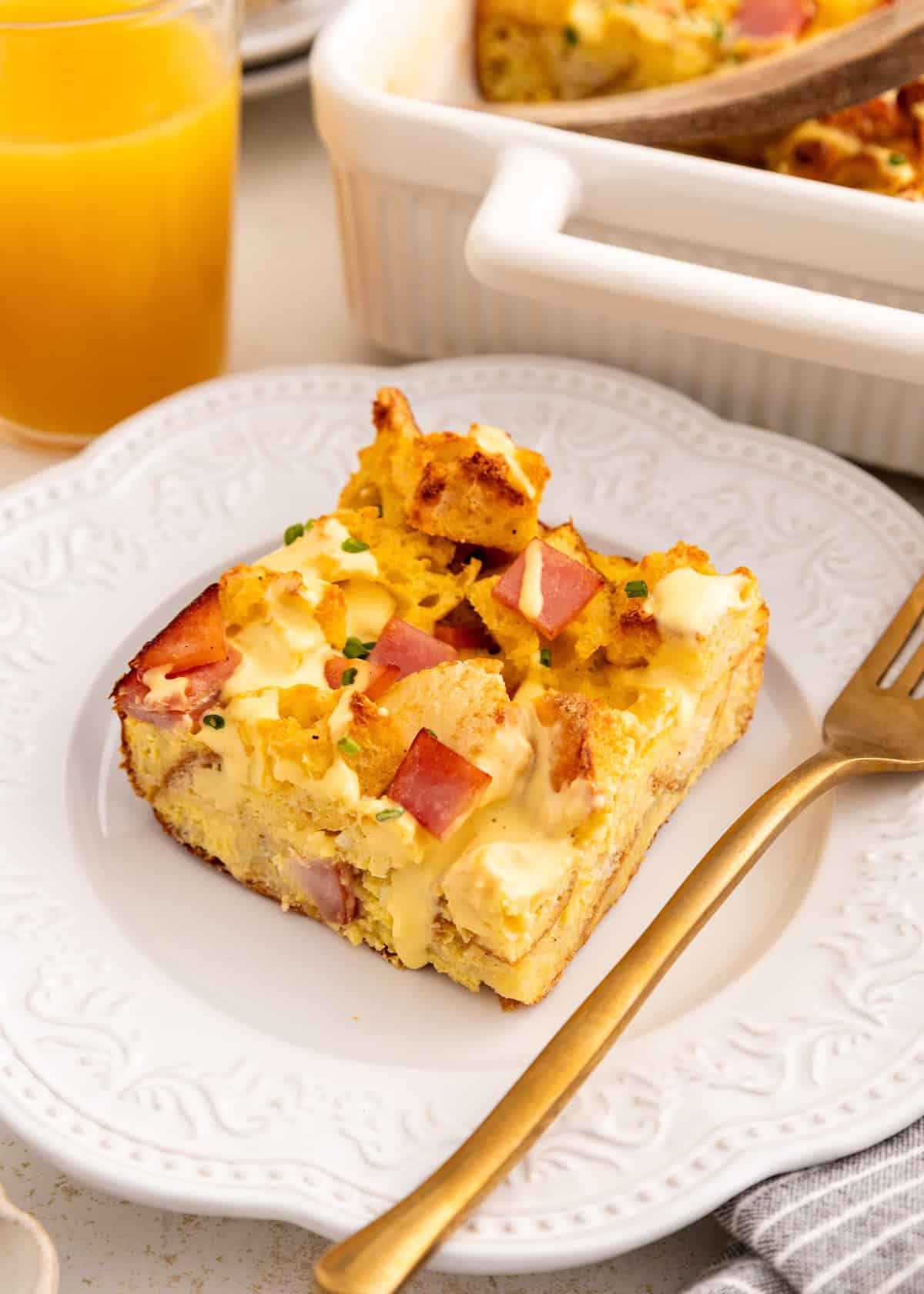 A slice of breakfast casserole with ham and cheese on a white plate next to a fork and orange juice.