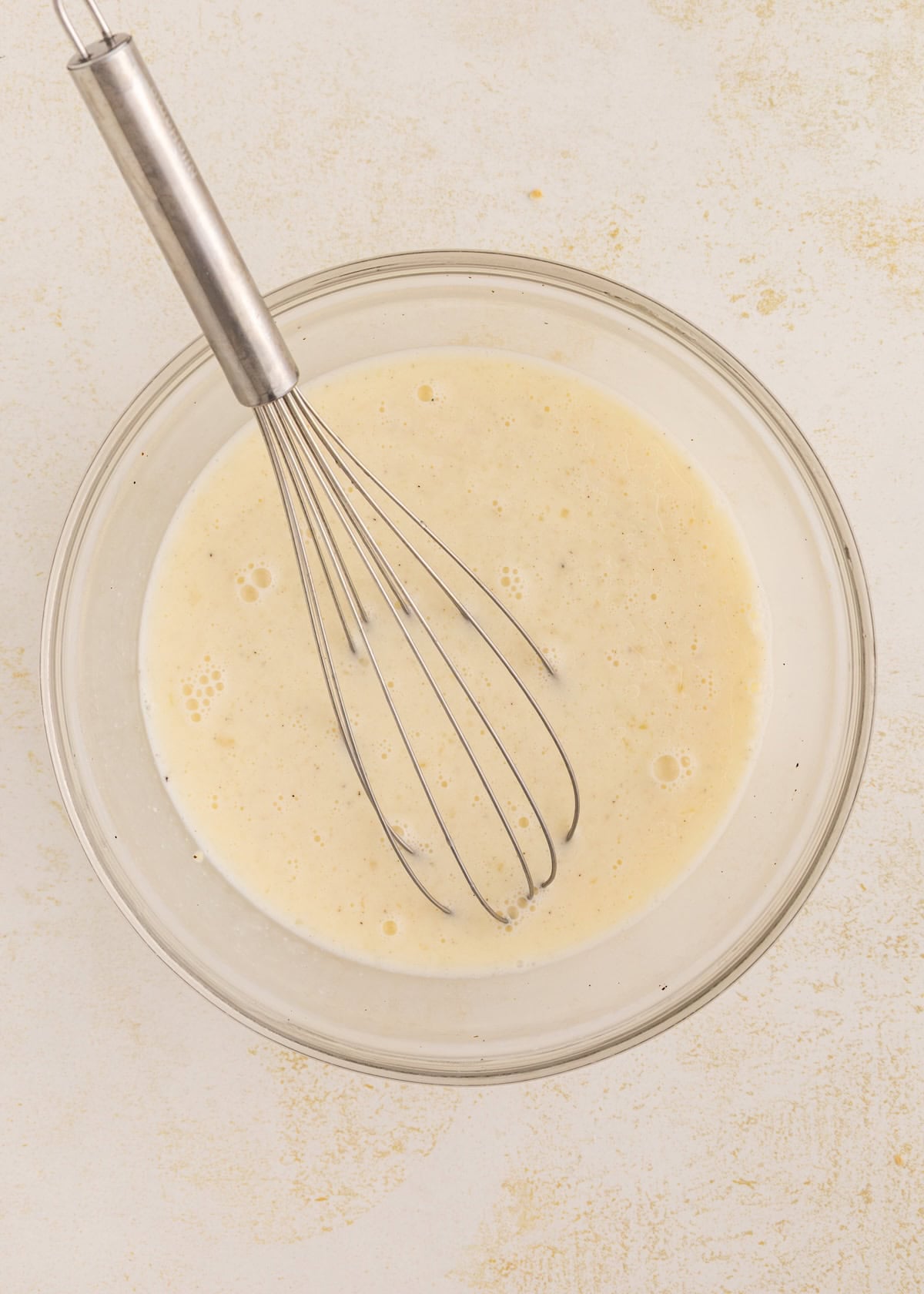 A metal whisk in a bowl of pale, creamy batter on a light surface.