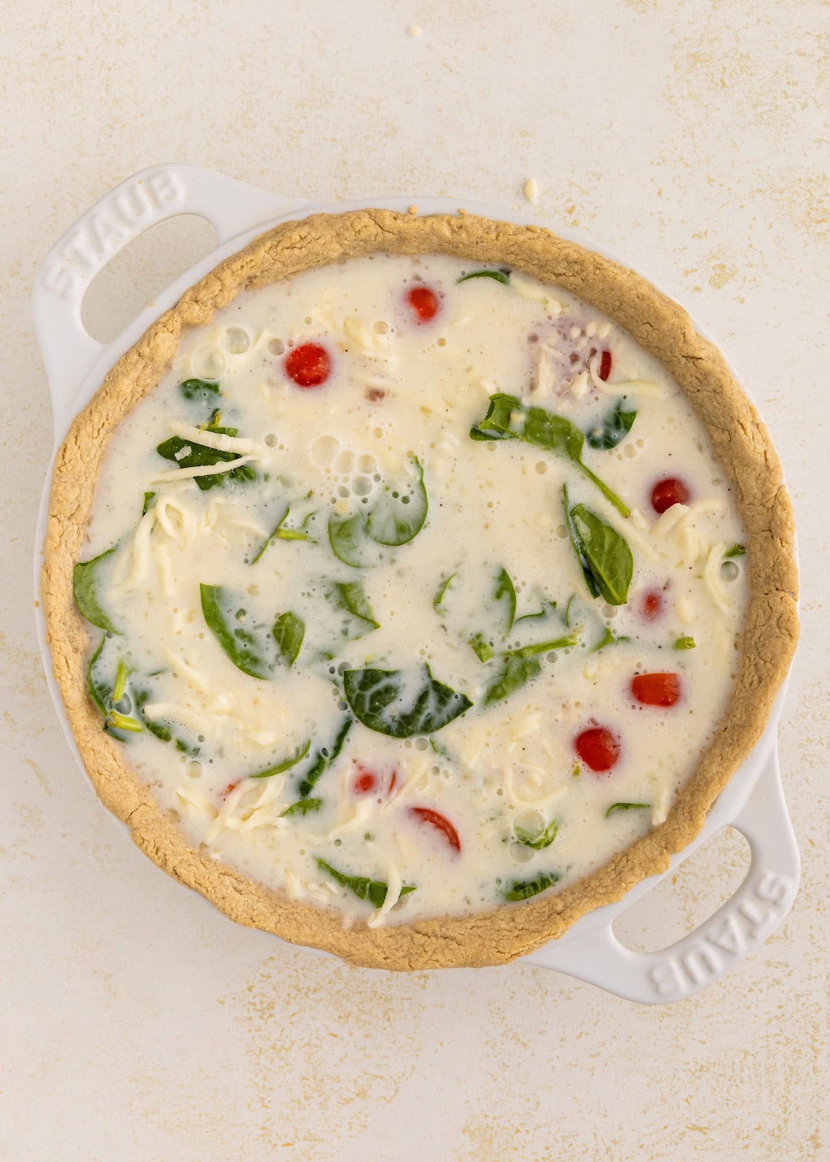 Unbaked quiche with spinach, tomatoes, and cheese in a round white baking dish.