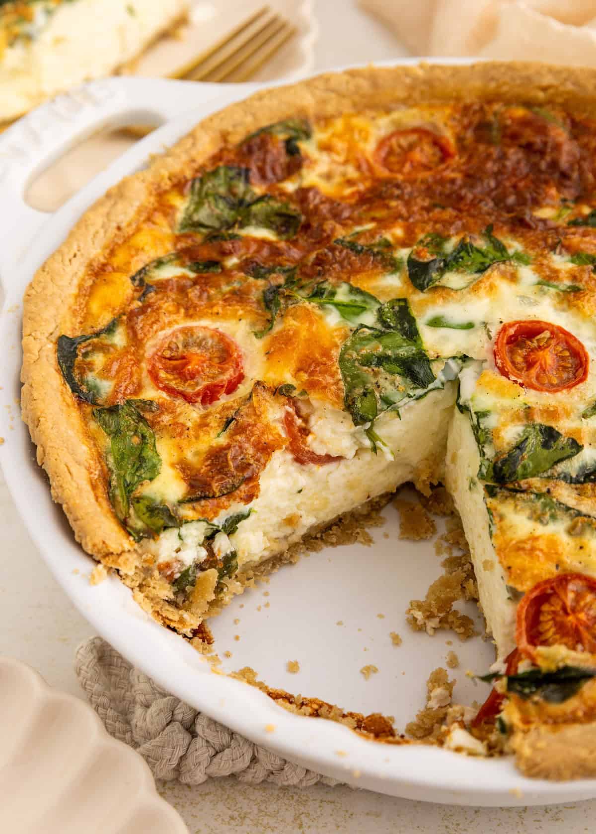 A spinach and tomato quiche with a slice missing in a white pie dish.