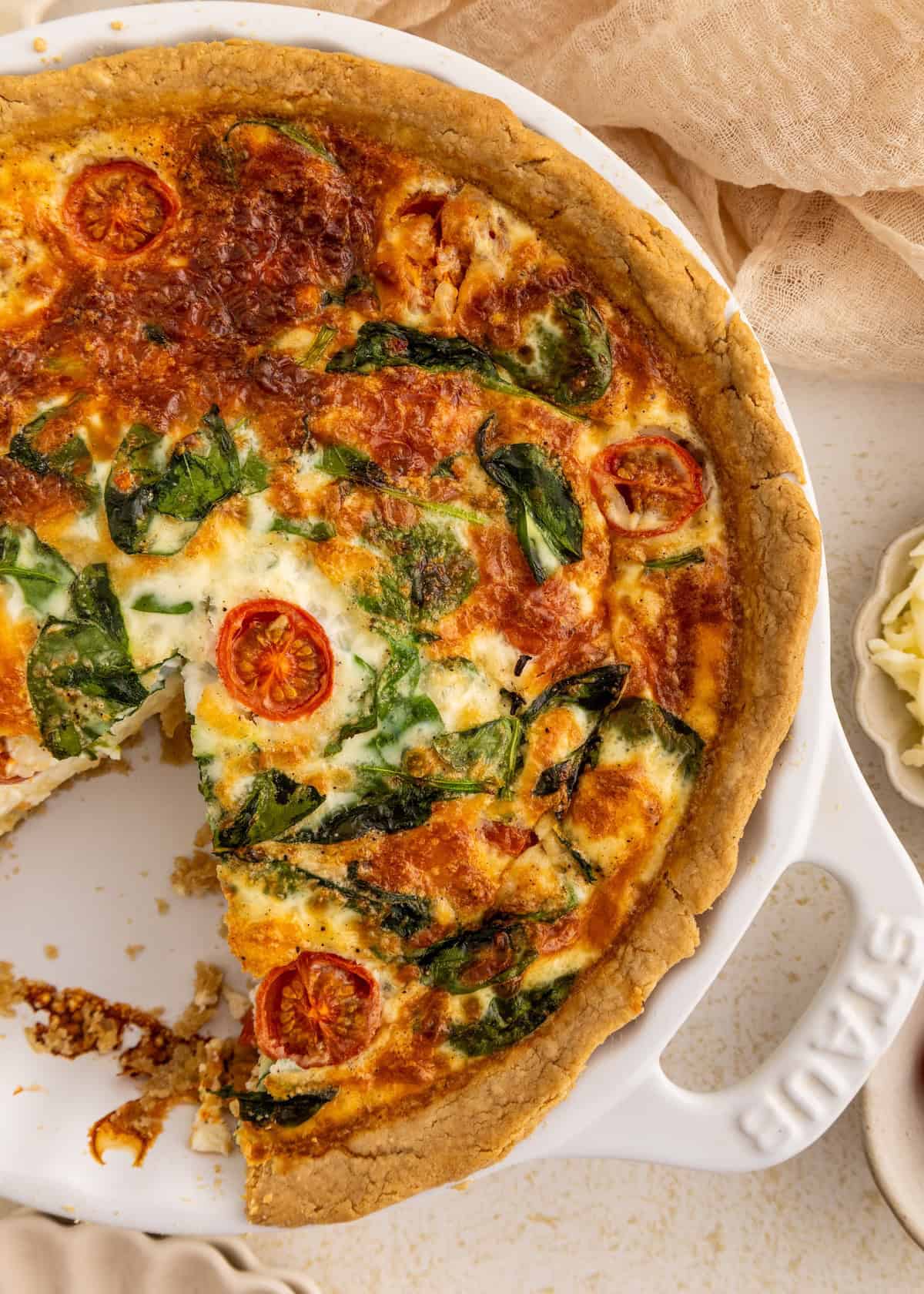 A baked quiche with spinach and cherry tomatoes, missing one slice, in a white dish.