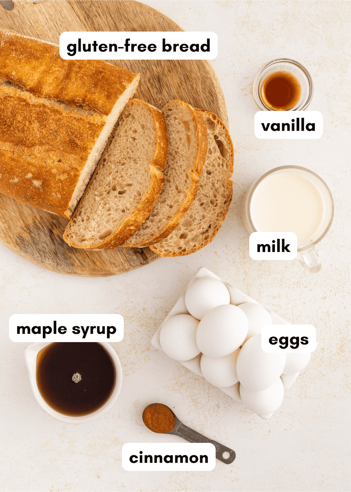 Sliced gluten-free bread, eggs, milk, maple syrup, vanilla, and cinnamon on a white surface.