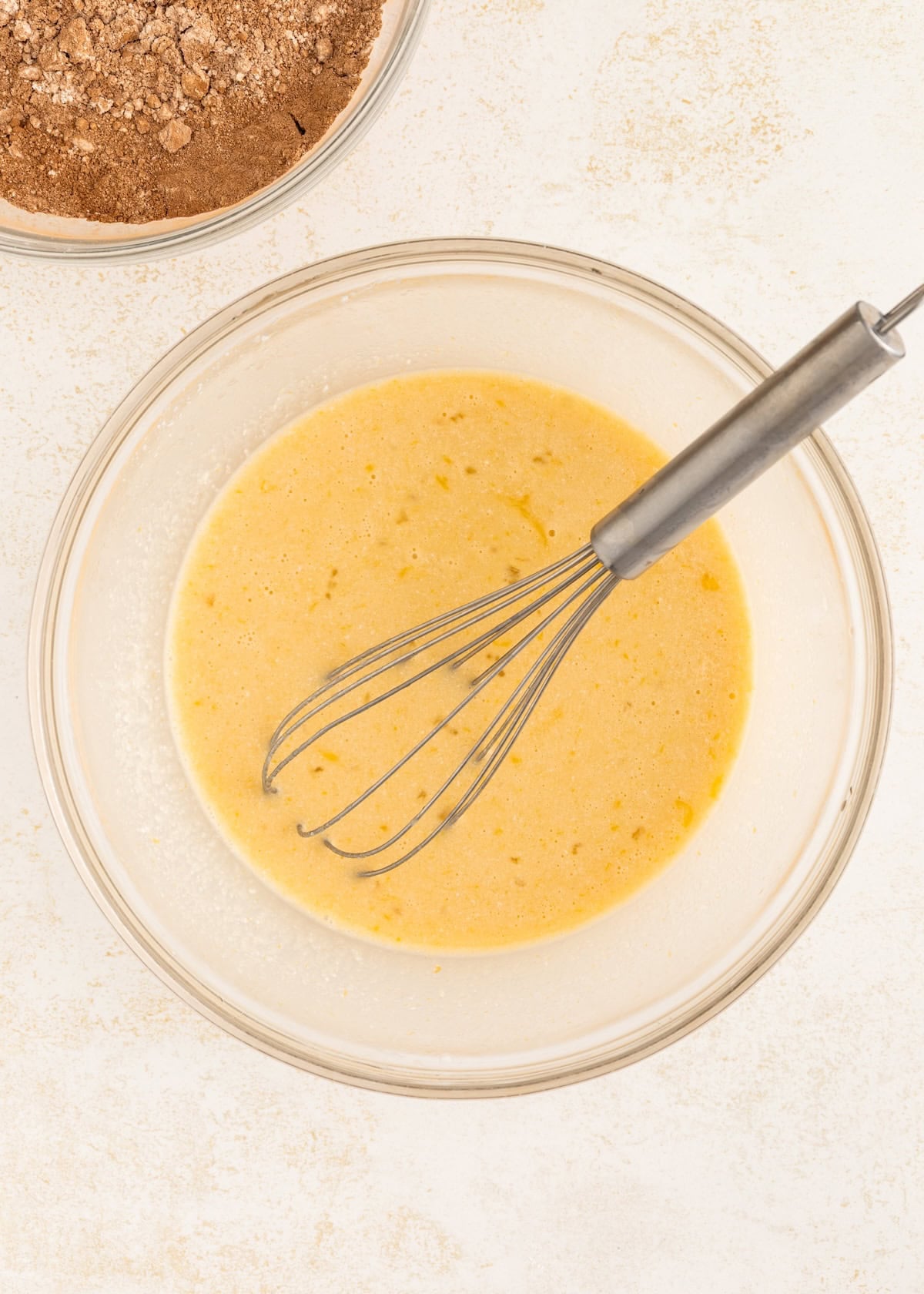 A metal whisk in a bowl of yellow batter, with a bowl of cocoa powder nearby.
