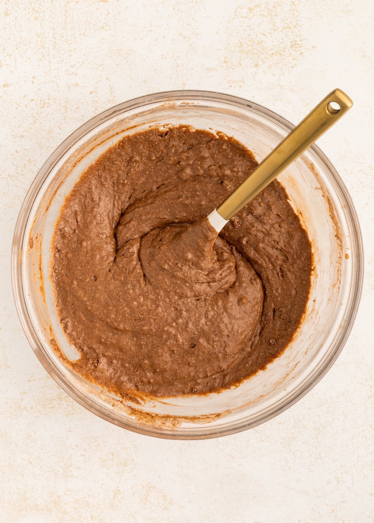 A glass bowl of chocolate cake batter with a gold spoon on a light surface.