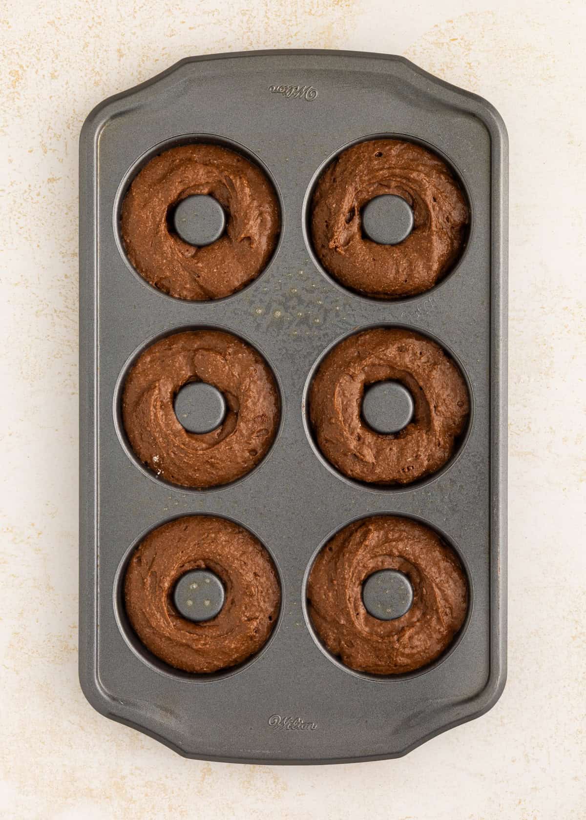 A metal donut pan filled with six unbaked chocolate donut batters.
