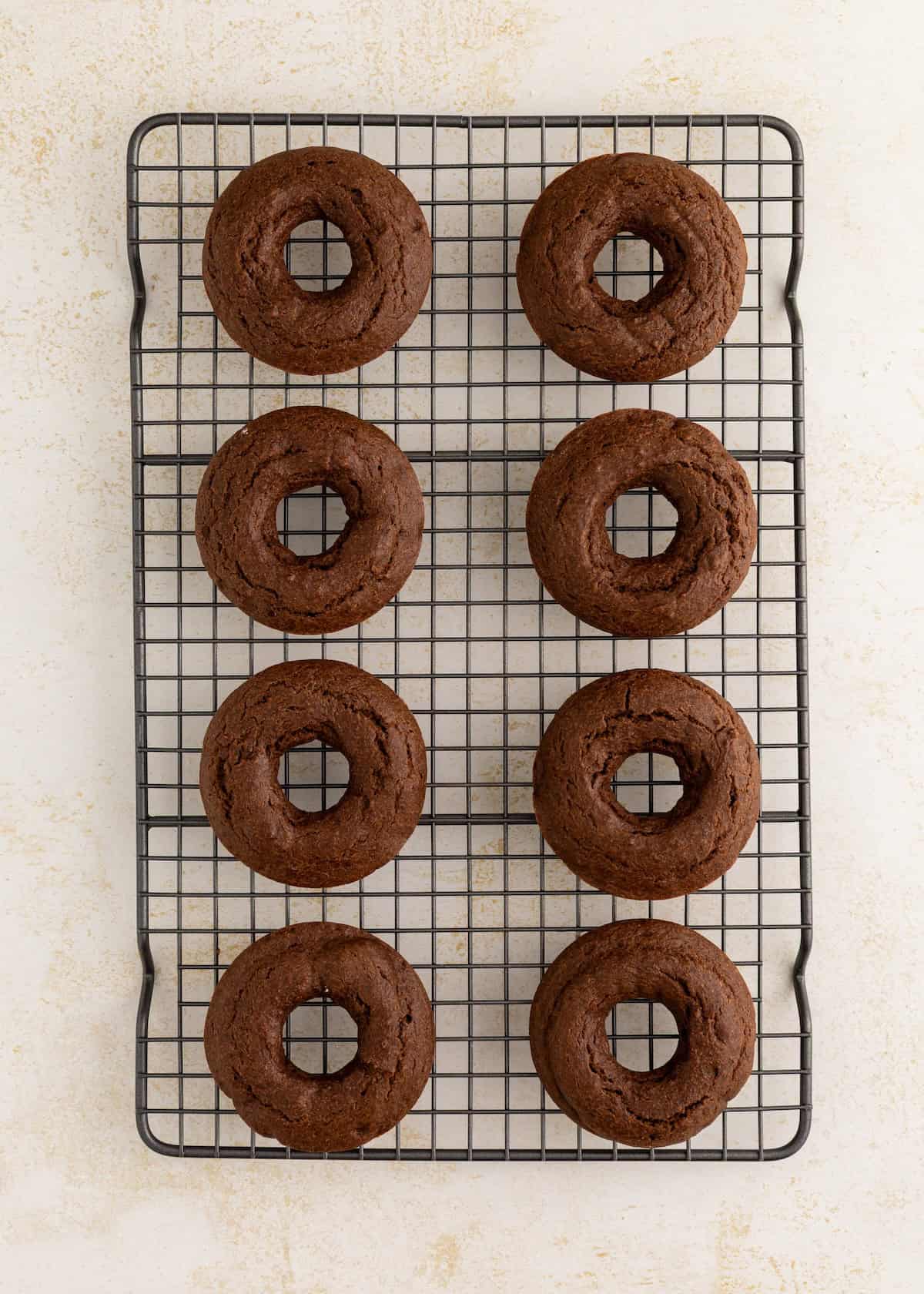 Eight chocolate donuts cooling on a wire rack against a light background.