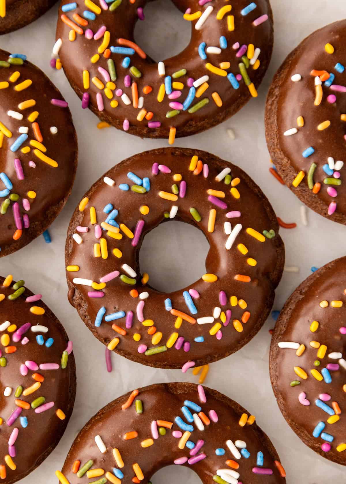 Chocolate donuts with colorful sprinkles arranged on a white surface.