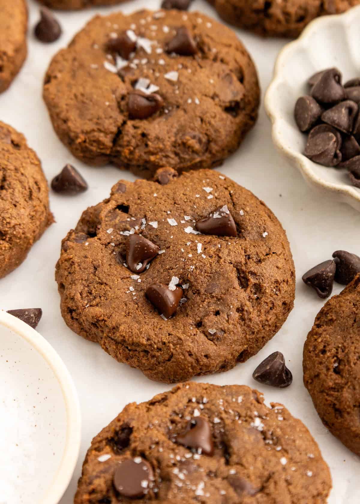 Chocolate chip cookies sprinkled with sea salt, surrounded by chocolate chips on a white surface.