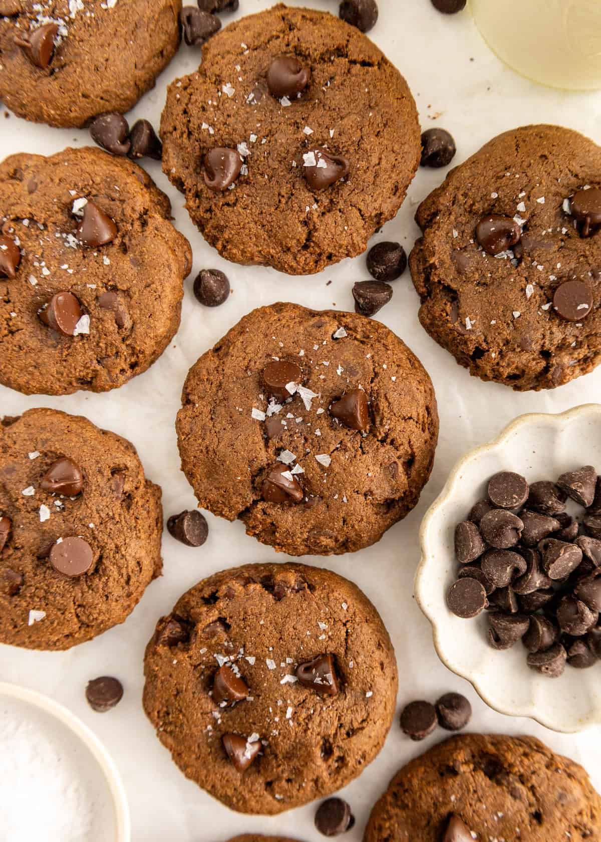 Chocolate chip cookies with sea salt on top, surrounded by chocolate chips on a white surface.